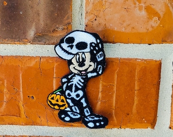 May include: A black and white skeleton Mickey Mouse costume patch with a pumpkin in hand. The patch is on a brick background.