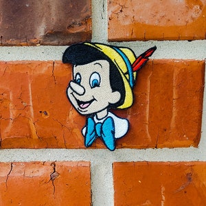 May include: A colorful embroidered patch featuring a cartoon character with a large nose, wearing a yellow hat with a red feather. The character has black hair, blue eyes, and a white shirt with a blue bow tie.