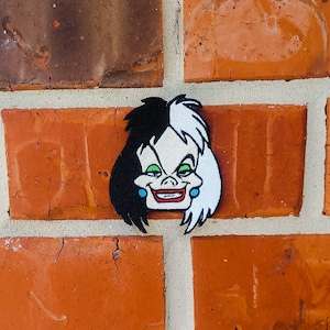 Puede incluir: Un parche bordado en blanco y negro de Cruella de Vil de la película de Disney 101 Dálmatas. El parche presenta su rostro con una sonrisa maliciosa y ojos verdes.