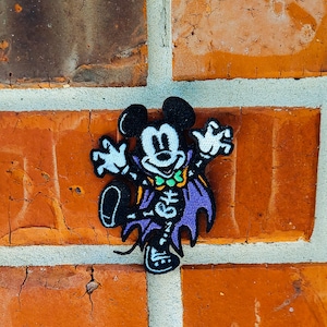 以下が含まれることがあります： ハロウィーンをテーマにしたパッチ。ミッキーマウスが吸血鬼のコスチュームを着ています。パッチは黒い背景に紫色のマント、スケルトンのディテール、緑の蝶ネクタイが付いています。パッチは赤いレンガの壁に取り付けられています。