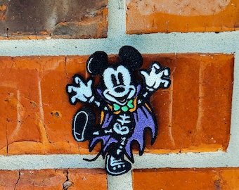 May include: A Halloween-themed patch featuring Mickey Mouse dressed as a vampire. The patch has a black background with a purple cape, skeleton details, and a green bow tie. The patch is attached to a red brick wall.
