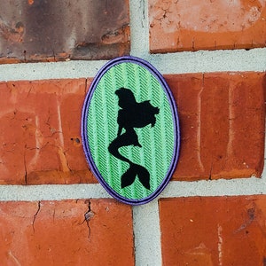 May include: A green and purple oval-shaped patch with a black silhouette of a mermaid sitting on a tail. The patch has a textured, striped pattern.