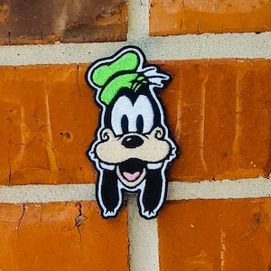 May include: A green, black, and white embroidered patch of Goofy, a cartoon dog character, with a green hat and a big smile.