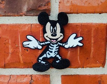May include: A black and white embroidered patch featuring Mickey Mouse in a skeleton costume. The patch has black ears, feet, and a skeleton design on the body. The background is a red brick wall.