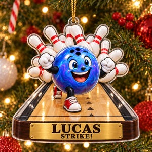 May include: A decorative Christmas ornament featuring a blue bowling ball character with arms and legs, standing on a wooden bowling lane with white pins in the background. A gold-colored plaque at the bottom displays the text LUCAS STRIKE!.