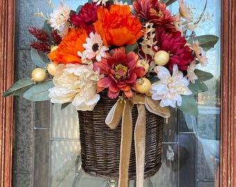 Könnte beinhalten: Dekorative Blumengesteck in einem Weidenkorb mit leuchtend orangefarbenen, roten und cremefarbenen Blumen. Der Korb ist mit goldenen Bändern verziert und wird von einem blau-weiß karierten Band aufgehängt, was einen einladenden Anblick erzeugt.