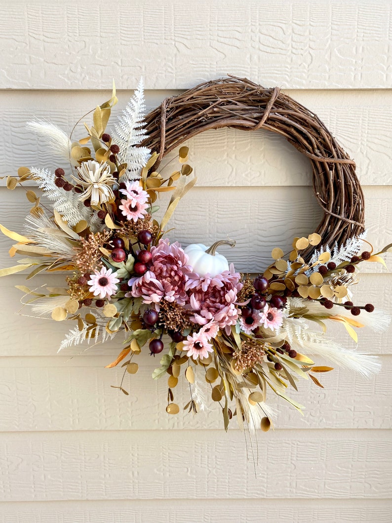 May include: A fall-themed wreath with a white pumpkin in the center. The wreath is made of grapevine and is decorated with dried flowers, pampas grass, and eucalyptus leaves in shades of gold, brown, and white. There are also pink flowers and burgundy berries on the wreath.