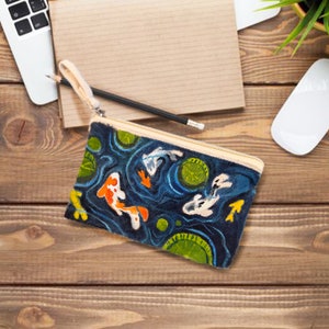May include: A rectangular, navy blue zippered pouch with a koi pond design. The pouch features orange, white, and yellow koi fish swimming among lily pads. The background is a dark blue with light blue swirls. A beige zipper and handle complete the design.