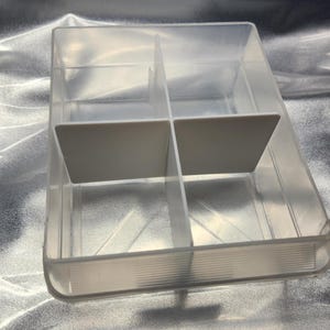 May include: A clear plastic storage box with four compartments. The box has dividers to separate the compartments.