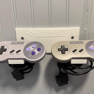 May include: A white wall-mounted Super Nintendo controller holder with two controllers. The controllers are gray with purple and black buttons. The holder has the Super Nintendo logo.