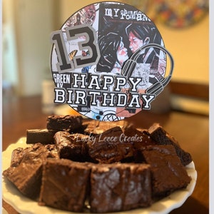May include: A round cake topper with a black and white design featuring the text "My Chemical Romance", a number "13", and the words "Happy Birthday". The topper is on top of a plate of chocolate brownies.
