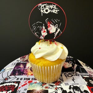 May include: A yellow cupcake with white frosting and colorful sprinkles, topped with a circular My Chemical Romance cake topper featuring a black and white illustration of two people with red accents. The cupcake sits on a collage of band imagery.