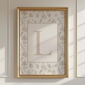 May include: A framed floral print with the letter "L" in a soft beige. The artwork has a cream-coloured background with a floral pattern, framed in a gold border. The print is displayed on a white wall, typical of home decor.