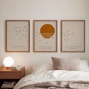 Op de afbeelding: Drie ingelijste kunstdrukken met een minimalistisch ontwerp. Elke print heeft een beige achtergrond, een zwerm zwarte vogels en tekst. Eén print heeft een oranje zongrafiek. De frames hebben een warme bruine kleur. De prints worden boven een bed weergegeven.