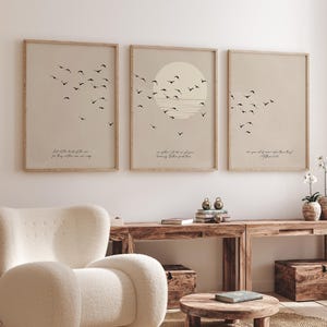 Könnte beinhalten: Drei gerahmte Kunstdrucke mit minimalistischem Design. Jeder Druck zeigt einen beigefarbenen Hintergrund, einen Schwarm schwarzer Vögel und ein Zitat in einer einfachen Schriftart. Der mittlere Druck zeigt eine Sonne, die über dem Horizont untergeht. Die Rahmen sind hellbraun.