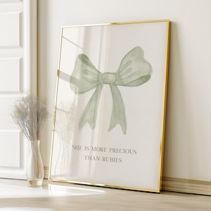 May include: A framed print with a gold border features a watercolor-style bow in shades of green. The text "SHE IS MORE PRECIOUS THAN RUBIES" is printed below the bow. The artwork is displayed on a light-colored wall.
