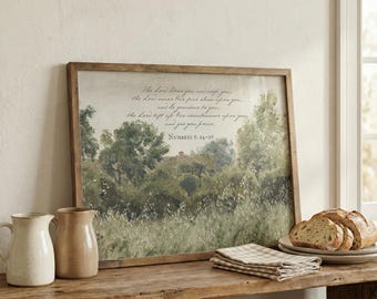 Könnte beinhalten: Ein gerahmter Landschaftsdruck mit Holzrahmen, der eine ländliche Szene mit Bäumen und Feldern zeigt. Der Text "The Lord bless you and keep you..." steht oben. Zwei Keramikkrüge und Brot liegen auf einem Holztisch.