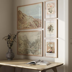 May include: Four framed prints on a white wall. The top left print is a landscape painting of a mountain range with a winding path. The top right print is a botanical illustration of a flower with the text 'Lovely Things' and 'September 1, 1883'. The bottom left print is a landscape painting of a river flowing through a valley with mountains in the background. The bottom right print is a botanical illustration of white lilies with the text 'Consider the Lilies' and 'September 1, 1883'. A wooden desk with a book and glasses on top is in the foreground.