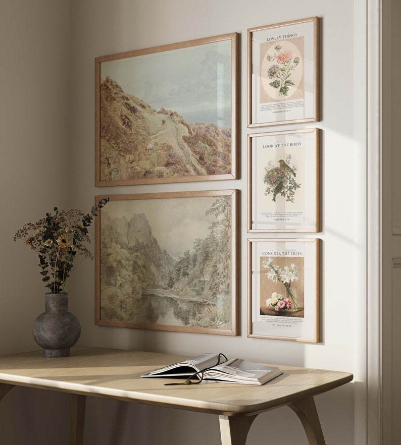 May include: Four framed prints on a white wall. The top left print is a landscape painting of a mountain range with a winding path. The top right print is a botanical illustration of a flower with the text 'Lovely Things' and 'September 1, 1883'. The bottom left print is a landscape painting of a river flowing through a valley with mountains in the background. The bottom right print is a botanical illustration of white lilies with the text 'Consider the Lilies' and 'September 1, 1883'. A wooden desk with a book and glasses on top is in the foreground.