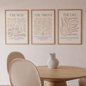 Puede incluir: Tres impresiones artísticas enmarcadas con diseños abstractos beige y crema y texto. Las impresiones dicen "THE WAY", "THE TRUTH" y "THE LIFE". Las impresiones se muestran encima de una mesa y sillas de madera, creando una estética minimalista.
