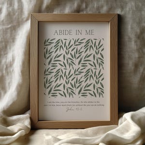 Op de afbeelding: Een ingelijste print met de woorden "ABIDE IN ME" bovenaan. De print heeft een patroon van groene bladeren op een beige achtergrond. Onder de bladeren staat een bijbelvers uit Johannes 15:5.