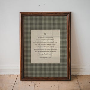 Puede incluir: Una impresión enmarcada con un marco de madera marrón oscuro. La impresión presenta un fondo beige con texto de Filipenses 4:6-7, sobre un patrón de cuadros verdes y grises. El texto está en una fuente serif clásica.