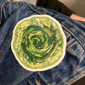 May include: A green and yellow embroidered patch of a swirling vortex, resembling the portal from the cartoon show "Adventure Time".
