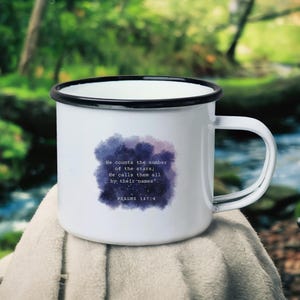 Könnte beinhalten: Eine weiße Emailletasse mit schwarzem Rand und Henkel. Die Tasse zeigt ein Aquarell-Design einer Galaxie mit dem Text "Er zählt die Zahl der Sterne; Er ruft sie alle mit ihren Namen." Psalm 147:4