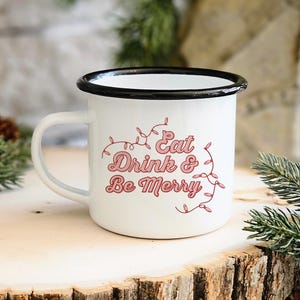 Peut inclure: Mug en émail blanc avec un bord et une anse noirs. La tasse porte l'inscription "Eat Drink & Be Merry" en script rose avec des guirlandes lumineuses décoratives. La tasse est posée sur une surface en bois avec des brins de conifères.
