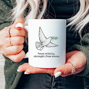 May include: White ceramic mug with a black and white illustration of a dove carrying a green olive branch. The mug has a red rim and the text "Peace within, strength from above."
