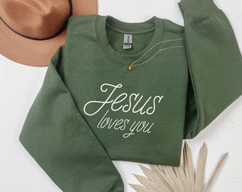 May include: An olive green sweatshirt with the phrase "Jesus loves you" in white script. A gold necklace with a small pendant and a brown hat are also visible. The sweatshirt is a casual, comfortable garment.