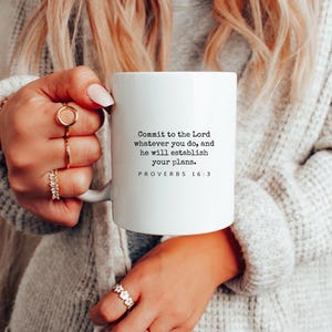 May include: White ceramic mug with the text "Commit to the Lord whatever you do, and he will establish your plans. PROVERBS 16:3." The mug is held by a person wearing rings and a sweater.