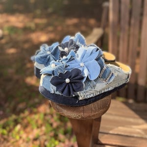 Könnte beinhalten: Jeansandalen mit Stoffblumen in Blau- und Weißtönen, akzentuiert mit Kunstperlen. Die Sandalen haben eine Used-Jeans-Optik mit dunkelblauem Besatz und hellblauem Grund. Die Blumen sind geschichtet und erzeugen einen strukturierten, dreidimensionalen Effekt.