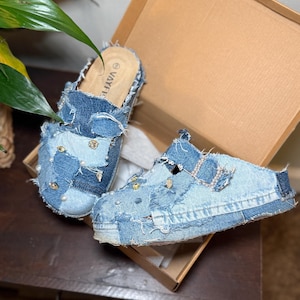 K&ouml;nnte beinhalten: Jeans-Mules mit Patchwork-Design. Die hellblauen Schuhe haben ausgefranste Kanten und sind mit Perlen und goldfarbenen Nieten verziert. Die Schuhe werden in einer Pappschachtel pr&auml;sentiert.