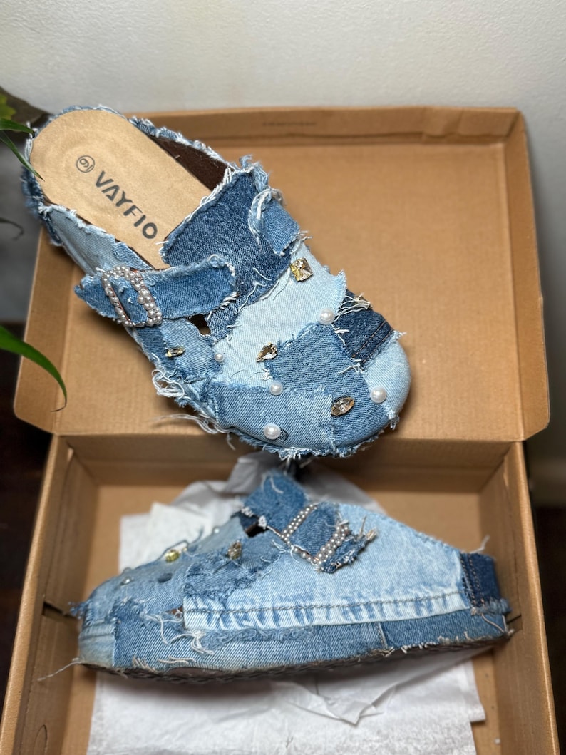 K&ouml;nnte beinhalten: Jeans-Clogs mit Patchwork-Design, verziert mit Perlen und Strasssteinen. Die Clogs haben einen Schnallenverschluss und befinden sich in einem Karton. Der Markenname "VAYFIO" ist auf der Innensohle sichtbar.