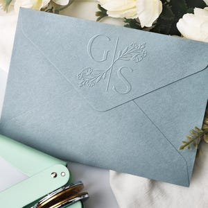 May include: A light blue envelope with embossed initials "G" and "S" intertwined with floral accents. A mint green envelope sealer is in the foreground. White flowers and greenery are in the background.
