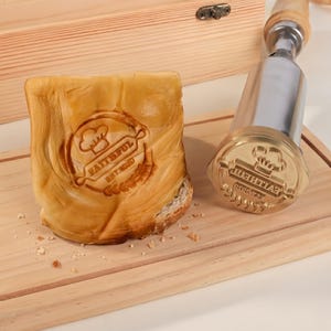 May include: A brass branding iron with a wooden handle featuring a chef hat, rolling pin, and the text FAITHFUL EST 2020, shown next to a piece of bread branded with the same design, resting on a wooden cutting board with a wooden box nearby.