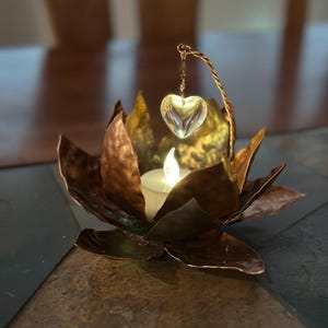 May include: A copper-colored lotus flower candle holder with a clear heart-shaped crystal hanging above a lit candle. The candle holder sits on a dark, textured surface. The overall design is elegant and decorative.