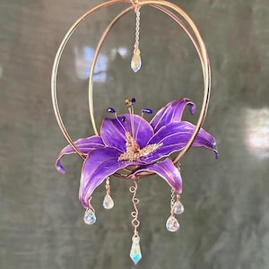 May include: A hanging ornament featuring a vibrant purple lily flower centred within two intersecting gold-toned metal rings. The flower has dark purple stamens and gold accents, with several clear, teardrop-shaped crystals suspended from the rings.