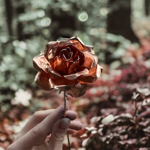 Puede incluir: Una rosa de metal color cobre con pétalos detallados se sostiene frente a un fondo natural borroso. El tallo de la rosa es delgado y oscuro, contrastando con la brillante flor metálica. La estética general es artística y elegante.