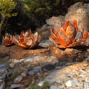 May include: Three copper lotus flower sculptures with a warm, metallic finish. The flowers are arranged on a natural, weathered wood base, set against a backdrop of rocks and greenery. The sculptures have a detailed petal design.