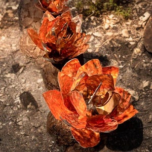 May include: Three copper-colored metal lotus flower sculptures. The flowers have detailed petals and are arranged on a rough, textured surface. The sculptures are in varying sizes, with the largest in the foreground.