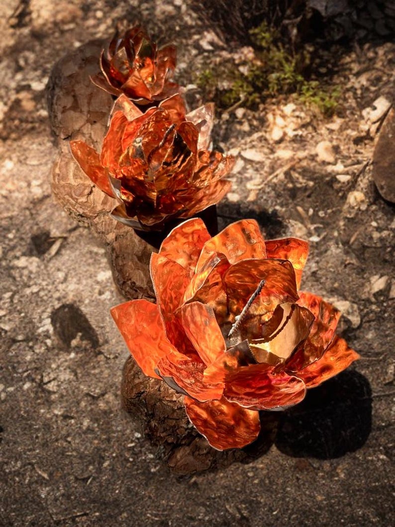 May include: Three copper-colored metal lotus flower sculptures. The flowers have detailed petals and are arranged on a rough, textured surface. The sculptures are in varying sizes, with the largest in the foreground.
