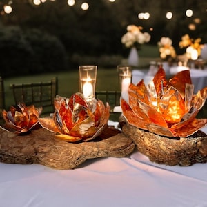 May include: Three copper-colored lotus flower candle holders on a wooden base. The candle holders are on a white tablecloth, with a blurred background of outdoor tables and string lights. The lotus flowers have a metallic finish.