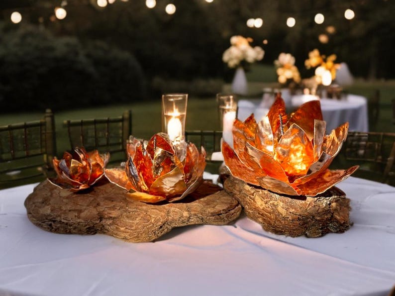 May include: Three copper-colored lotus flower candle holders on a wooden base. The candle holders are on a white tablecloth, with a blurred background of outdoor tables and string lights. The lotus flowers have a metallic finish.