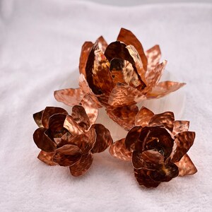 May include: Three copper-colored metal lotus flower sculptures. The flowers have textured petals and are arranged on a white surface. The largest flower is in the background, with two smaller flowers in front.
