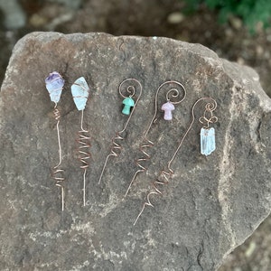 May include: A collection of copper wire plant stakes with various crystal and gemstone toppers. The toppers include amethyst, clear quartz, green and pink mushroom shapes, and a clear crystal. The stakes are arranged on a gray stone surface.