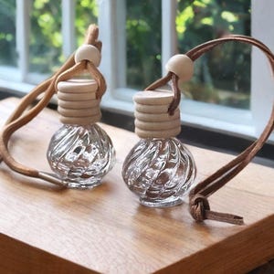 Puede incluir: Dos frascos de aromaterapia de vidrio transparente con tapas de madera y cordones de cuero marrón. Los frascos tienen un diseño texturizado y arremolinado. Los cordones están atados con un nudo y una cuenta de madera. Sobre una superficie de madera.