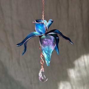 May include: A handcrafted, blue and purple flower-shaped ornament with a copper chain and a clear crystal teardrop. The flower has a metallic finish and is suspended in the air, creating a whimsical and decorative piece.