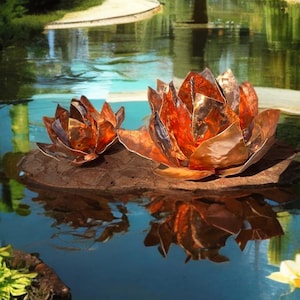 Könnte beinhalten: Zwei Kupfer-Lotusblumen-Skulpturen, die auf einem Teich schwimmen. Die Blumen haben komplizierte Blütenblattdesigns und einen warmen, metallisch-orangen Farbton. Der Teich spiegelt die Blumen und das umliegende Grün wider.